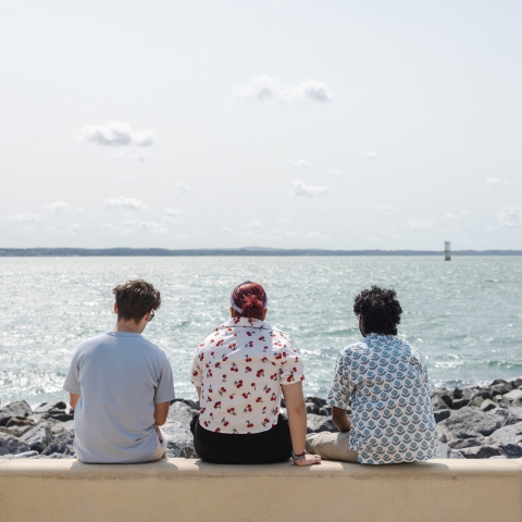 Three students sat on a wall by the sea, backs to the camera