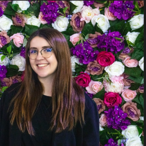 Creative Writing and Film Industries graduate, Mollie Winter, smiling in front of a colourful flower wall.