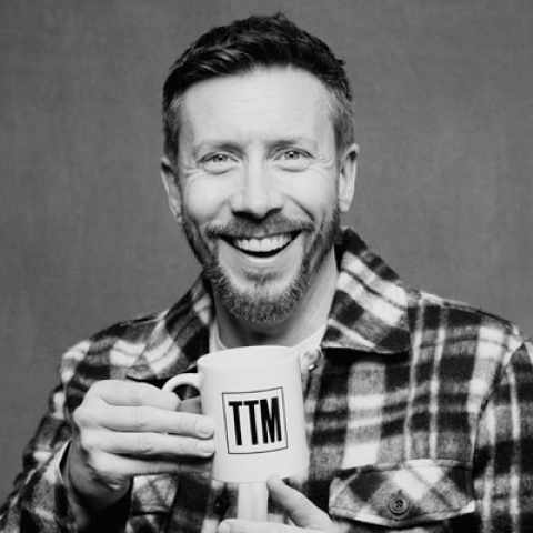 Headshot of Sam Matterface smiling to camera wearing chequered shirt and holding up a mug to camera