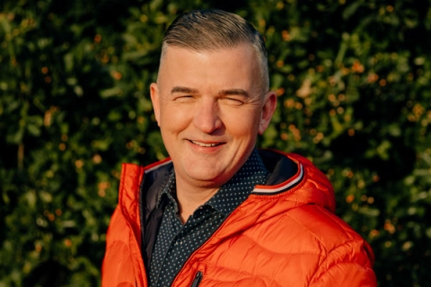 Headshot of Craig Jones MBE wearing orange jacket and smiling to camera with greenery backdrop