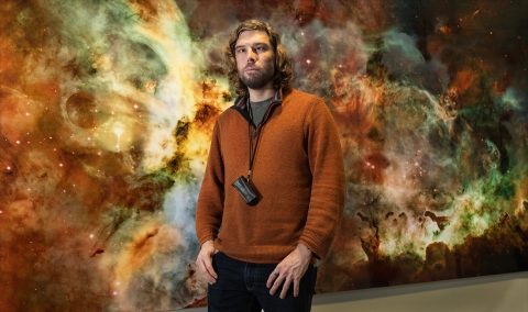 Dr Nicolas Bonne against a backdrop featuring a Hubble Space Telescope image of a small section  of the Carina Nebula - a vast cloud of dust and gas that contains  thousands of stars