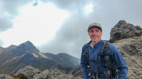 A man with a backpack and blue shirt stands on a mountain enjoying the panoramic view around him.