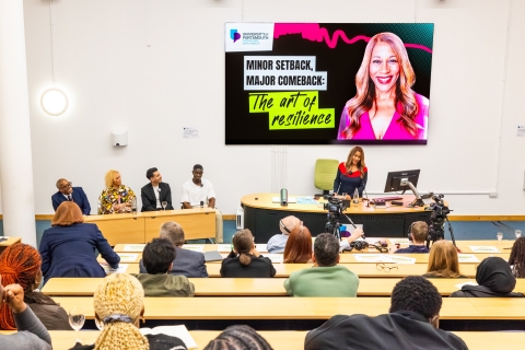 A lecture theatre with rows of students facing Karen Blackett speaking at a lectern and a panel of spekers
