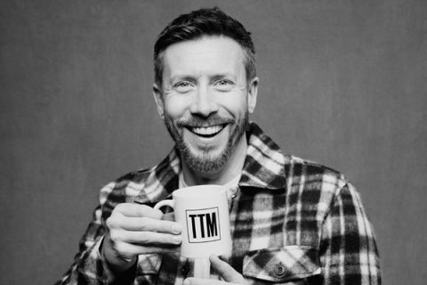 Headshot of Sam Matterface smiling to camera wearing chequered shirt and holding up a mug to camera