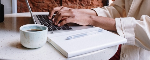 Person in white long sleeve shirt using a laptop with a notebook and cup of coffee
