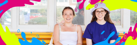 Two students sat at the beach, one wears a hat. 