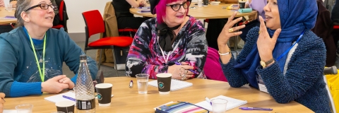 A group of women seated around a table engaged in a lively discussion at a professional event. One woman in a blue hijab is speaking animatedly, using hand gestures, while two others, one wearing glasses and a blue sweater and the other with vibrant pink hair and a patterned outfit, listen attentively with smiles. The table is filled with notebooks, coffee cups, and event materials. In the background, other attendees are seen networking and conversing.