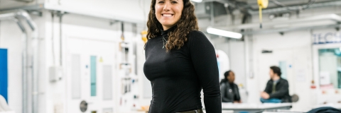 A person with long hair standing in front of a worktable, smiling to camera