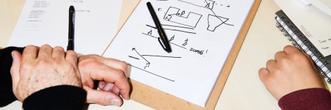 A clipboard, notes and pen on a table, with two sets of hands visible.