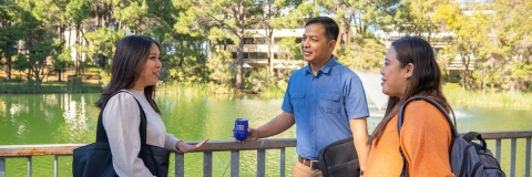 three people talking on a bridge in front of a lake