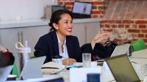 A student sat at a table wearing a black blazer talking to other students, smiling 