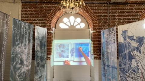 A picture of the Site Lines and Motifs exhibition. In the centre is a screen with some artwork and to either side are fabric printed banners