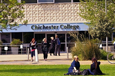 A view of the outside of the main entrance to Chichester College with students walking and sitting on the grass