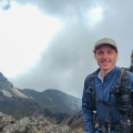 A man with a backpack and blue shirt stands on a mountain enjoying the panoramic view around him.
