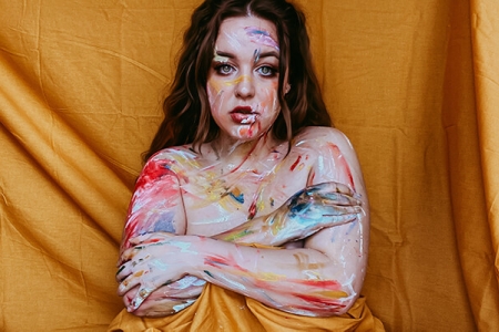 Profile image of a naked lady painted with different colours hiding herself with the background orange fabric