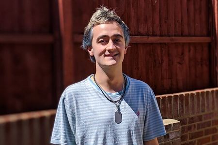 A male person wearing glasses in a grey top, with a pendant round his neck, standing by a fence
